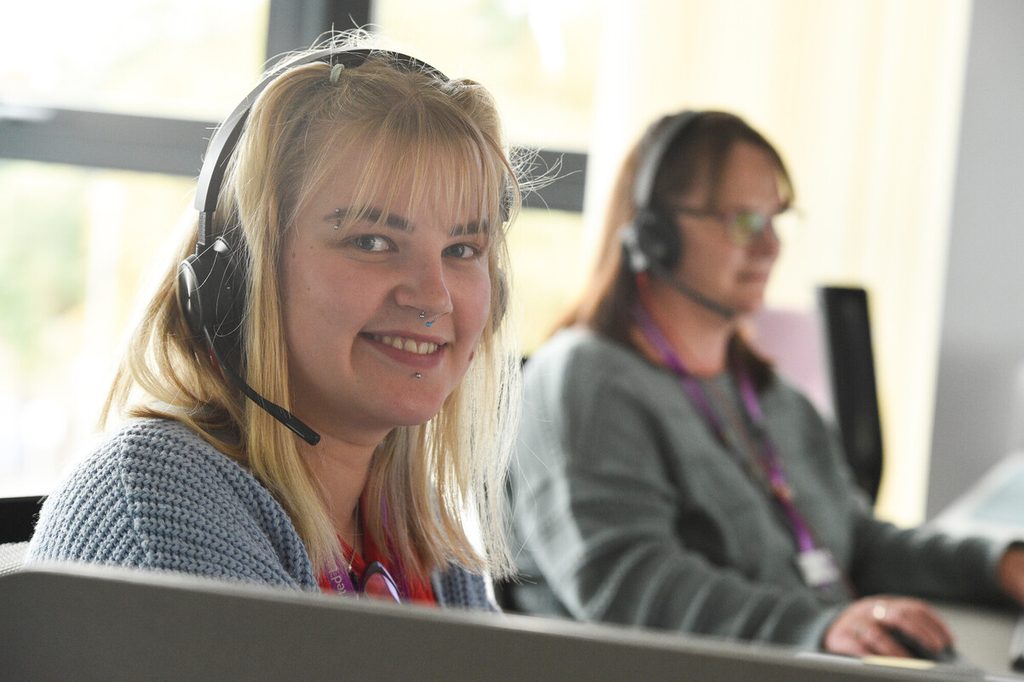 A young woman with blonde hair and a friendly smile is wearing a headset and sitting at a desk in an office environment. She has facial piercings and is looking directly at the camera. In the background, another woman with brown hair, also wearing a headset, is focused on her work. The setting is bright and modern, with large windows allowing natural light to enter.