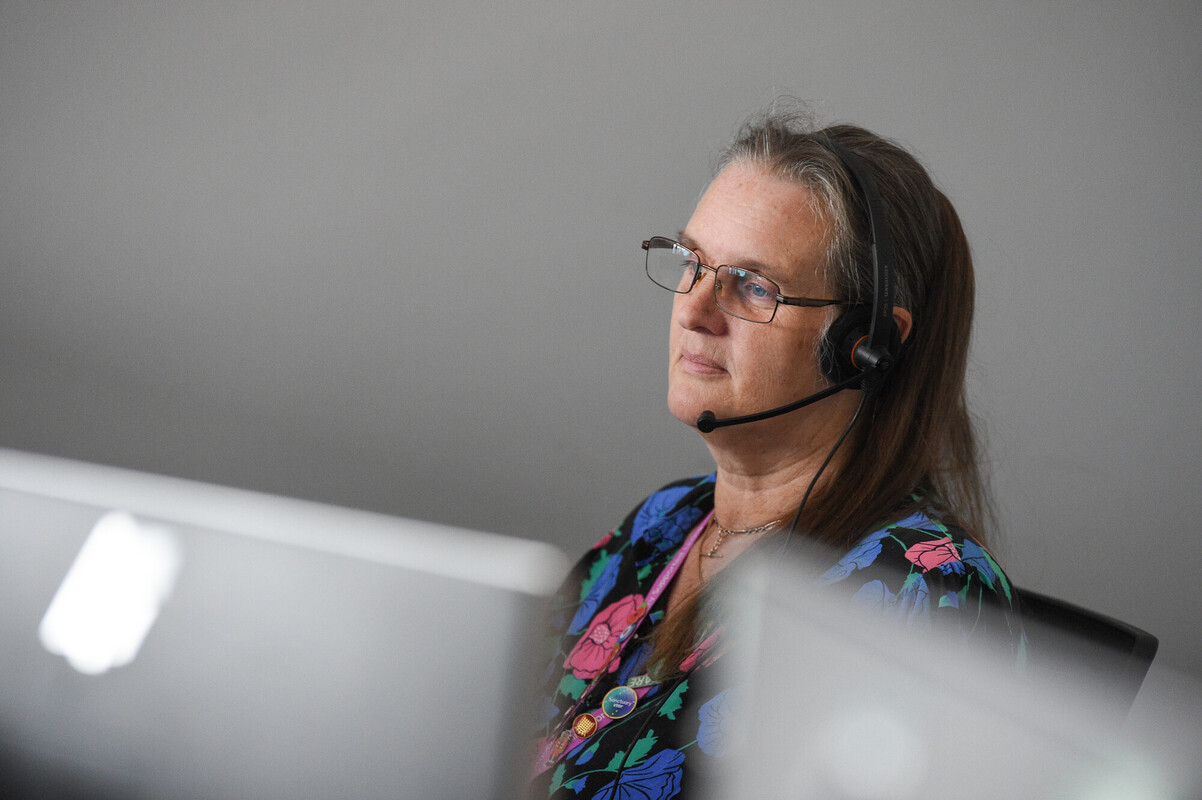 A woman with long hair and glasses is sitting at a desk, wearing a headset. She has a thoughtful expression and is dressed in a colorful floral blouse. In the background, there are blurred computer screens, suggesting a work environment.