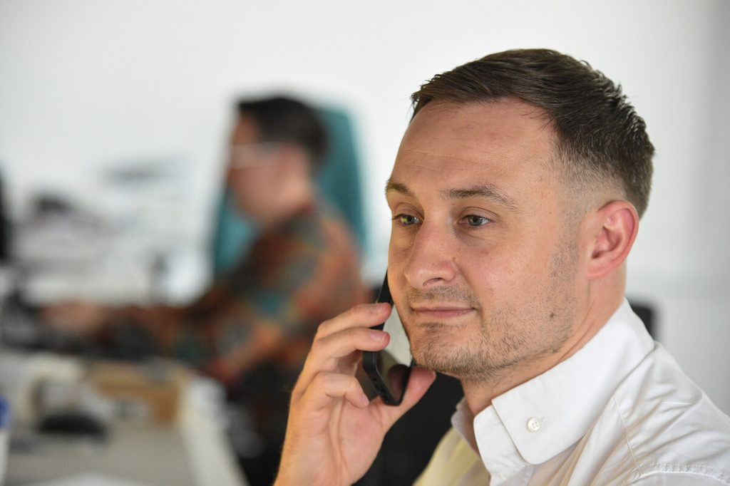 A man with short hair and a slight smile is holding a smartphone to his ear, engaged in a conversation. He is wearing a white shirt and appears focused. In the background, another person is working at a desk, partially visible, with a blurred colorful shirt. The setting is an office environment with modern decor.